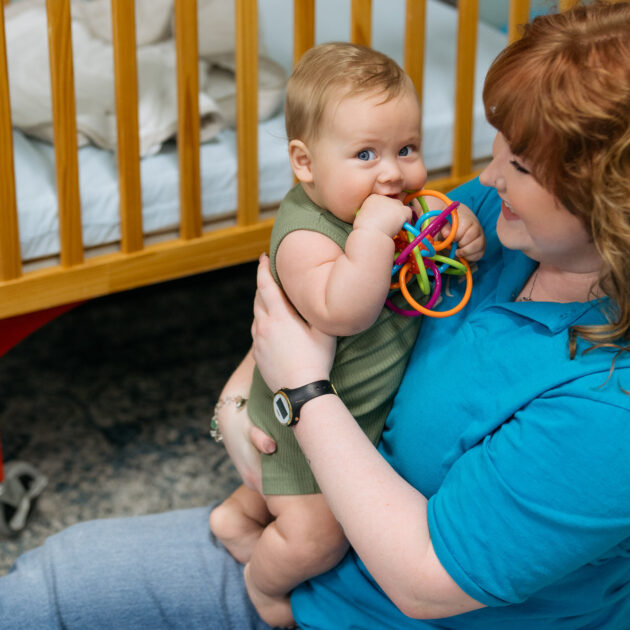 A woman in a blue shirt holds a baby in a green outfit who is chewing on a colorful plastic ring toy near a wooden crib.