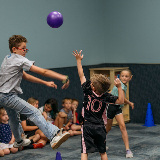 Three children actively play with a purple ball in a room while other kids sit on the floor against the wall watching. Blue cones mark areas on the floor.