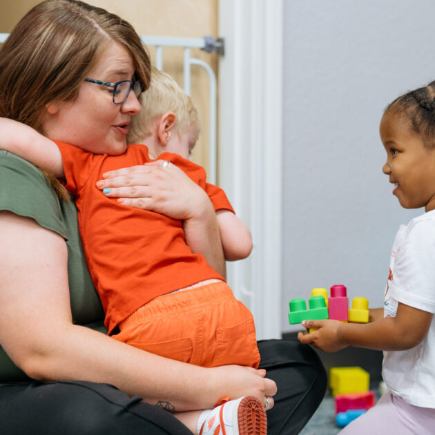 A woman comforts a child in her lap while another child holding toy blocks smiles nearby; a fourth child plays in the background.