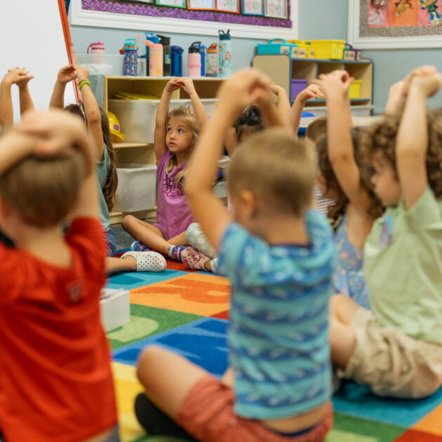 Young children sit in a circle on a colorful classroom rug, raising their arms above their heads, participating in a group activity.