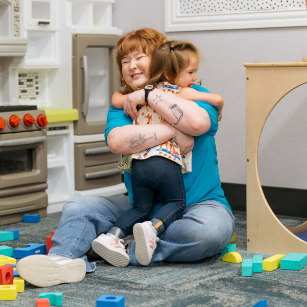 An adult and a child sit on the floor of a playroom, hugging each other amid scattered colorful plastic blocks and play kitchen furniture.