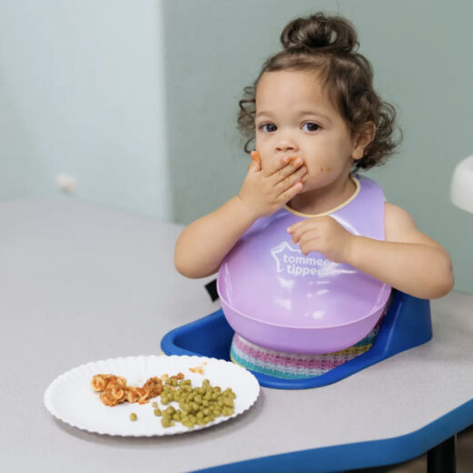 A toddler wearing a purple bib sits at a table, eating pasta and peas from a paper plate, with food on their face and hand.