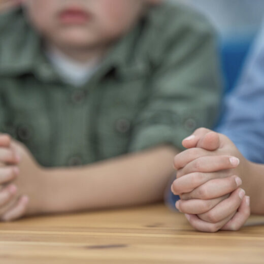 Two children sit at a table with their hands clasped together, shown from the shoulders down. Their faces are not fully visible.