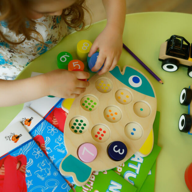 A child plays with a wooden fish-shaped puzzle with numbered pieces on a table, next to toy trucks, pencils, and colorful books.