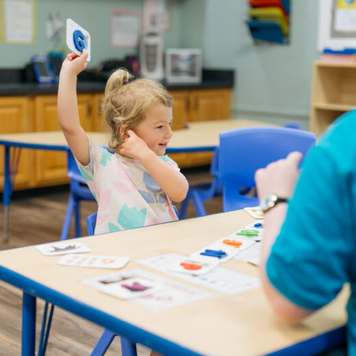 A young girl sits at a classroom table raising a card in her hand, participating in an activity with an adult across from her.